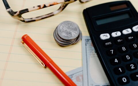 Close-up of a workspace with a calculator, coins, and glasses on a notepad.