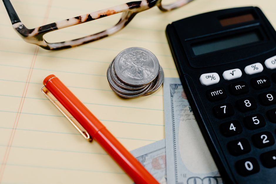 Close-up of a workspace with a calculator, coins, and glasses on a notepad.