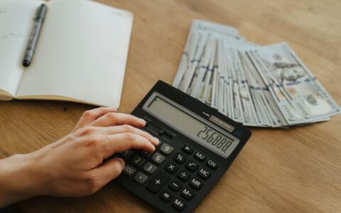 A hand calculates financial figures using a calculator with stacks of cash nearby on a wooden table.