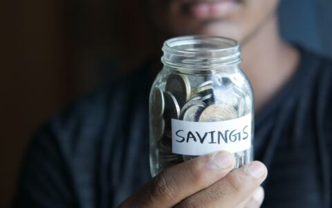A close-up image of a person's hand holding a jar full of coins labeled 'Savings'.