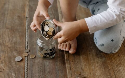 A child adds coins into a glass jar labeled for savings on a wooden floor.