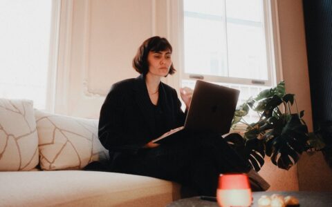 Woman in suit using laptop on sofa in bright room, modern lifestyle.