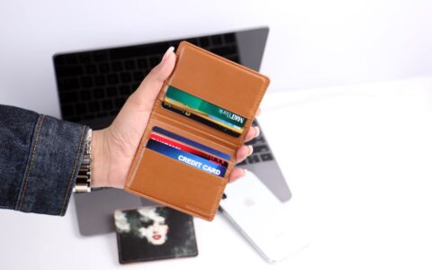 Close-up of a hand holding a leather wallet with credit cards, indoors with a laptop background.