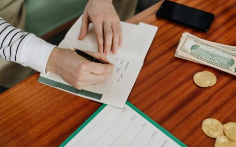 Person writing in a notebook on a wooden table with cash, coins, and a phone nearby.