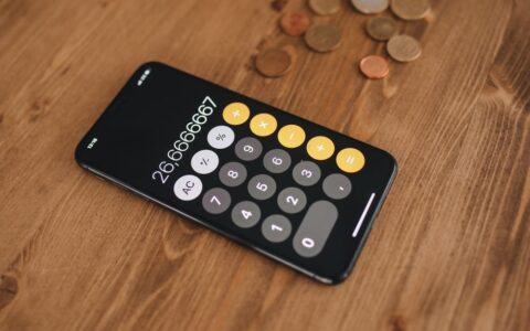 A close-up of a smartphone displaying a calculator app surrounded by coins on a wooden table.