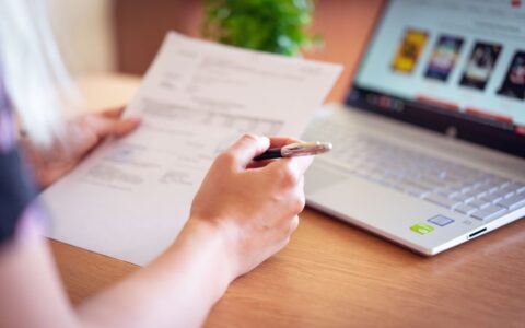 Close-up of a hand holding a pen and paper near a laptop, suggesting online shopping or document review.