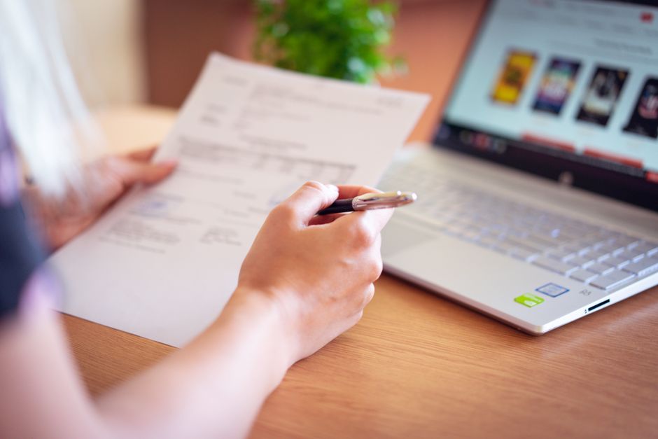 Close-up of a hand holding a pen and paper near a laptop, suggesting online shopping or document review.