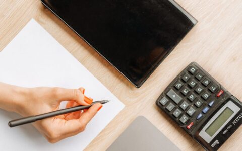 Top view of a minimalist office desk with a hand writing, calculator, and electronic device.