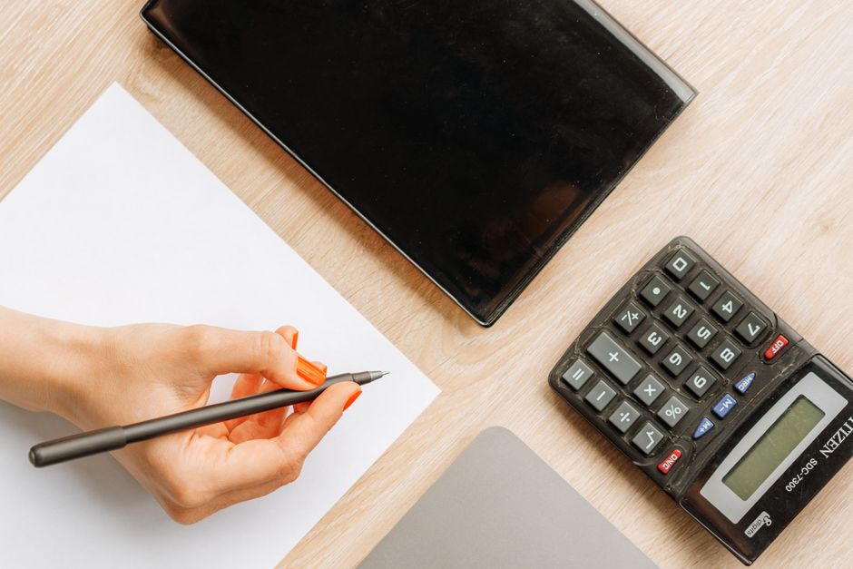 Top view of a minimalist office desk with a hand writing, calculator, and electronic device.