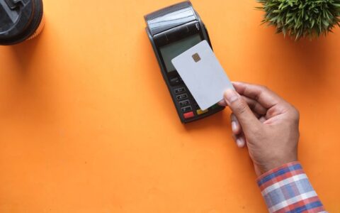 A person making a contactless payment with a credit card and card reader on a bright orange surface.