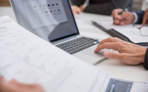 Close-up of hands working on documents and a laptop in an office setting, illustrating teamwork and productivity.