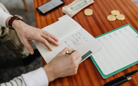 Hands writing financial calculations on notebook with money and coins on table.